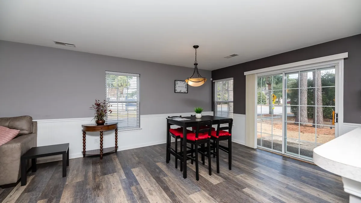 A dining area with a table and red chairs, adjacent to a sofa and large windows streaming in natural light. The space is arranged for both casual and special gatherings, creating a seamless flow between dining and living areas.