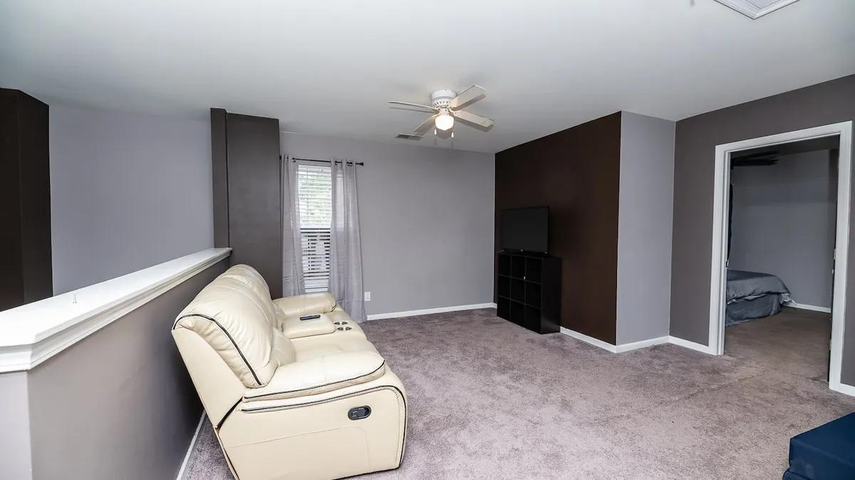 A cozy living room featuring a beige recliner sofa, a television unit, and a window with curtains. The setup is designed for relaxation, socializing, and casual entertainment—perfect for unwinding after exploring the city.