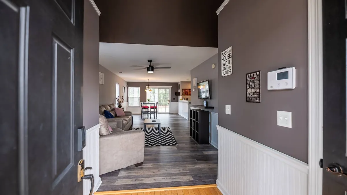 This image captures a spacious, airy living area with a comfortable sofa, a visible dining space, and a television. Large windows and light color tones create a bright, inviting atmosphere ideal for socializing, relaxing, or sharing meals. The open sightlines between the living and dining areas highlight the open-concept design.
