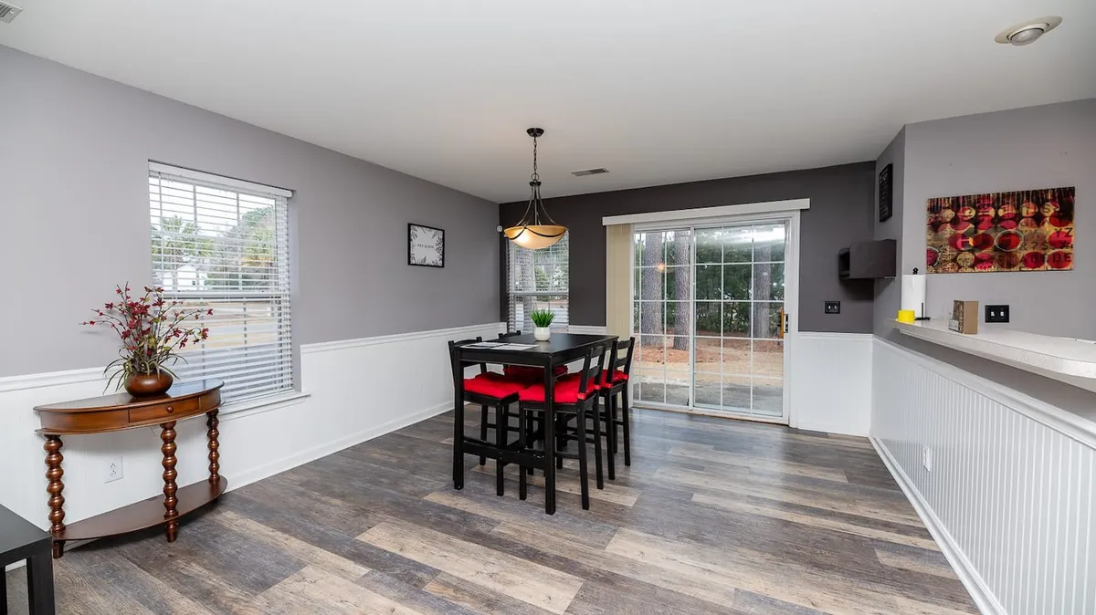 A dining area with a spacious table surrounded by chairs, a window letting in natural light, and a decorative plant. The setup encourages group meals, coffee breaks, and festive gatherings.