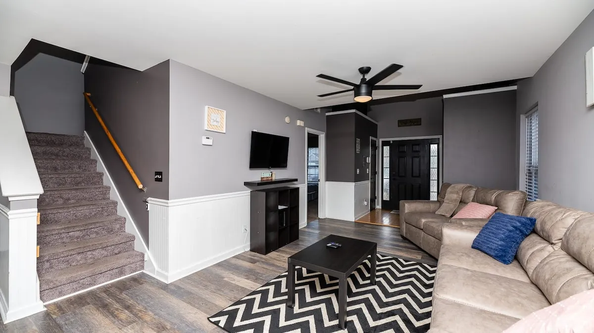 A cozy upstairs loft space featuring a comfortable sofa, a playful zigzag rug, a television, and a visible staircase. The atmosphere invites guests to relax, watch movies, socialize, or read, with plenty of open floor space to accommodate kids' play or group activities.