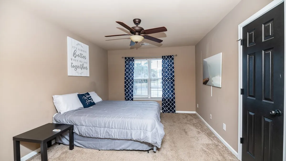 A softly lit bedroom with a comfortable bed positioned near a large window, inviting natural light into the space. Tasteful wall decor and a nightstand complete the scene, while a ceiling fan hints at gentle comfort. The atmosphere is calm and tranquil, perfect for relaxing or reading.