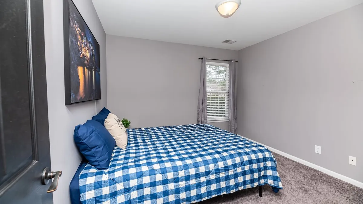 A calming bedroom outfitted with a blue checkered quilt, framed artwork on the wall, and a window that fills the room with natural light. The overall mood is restful and bright, ideal for waking up refreshed or enjoying a quiet moment.