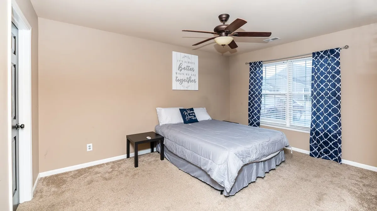 A serene bedroom with a neatly made bed, window with curtains allowing in soft daylight, a decorative sign, and gentle, calming colors. The space feels peaceful, inviting guests to enjoy both privacy and comfort.