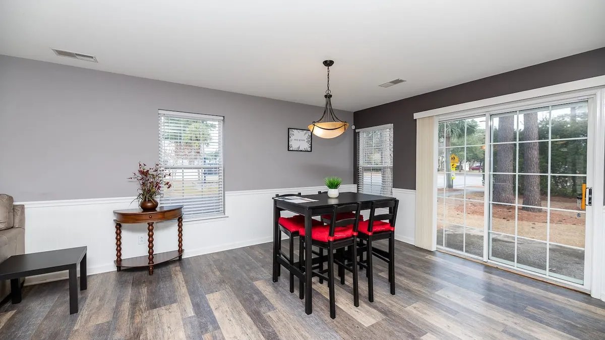 A bright, inviting dining area with a large wooden table surrounded by bold red chairs, bathed in natural light from a wide glass door that opens to the outdoors. A tasteful flower arrangement sits on a side table beneath a classic wall clock, creating a space that feels both homey and elegant.
